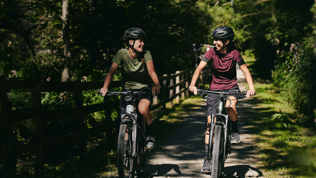 Touring bikes combine durability and comfort for your long-distance adventures. Two women riding Canyon e-touring bikes through a scenic, wooded countryside trail, enjoying a casual ride in nature.