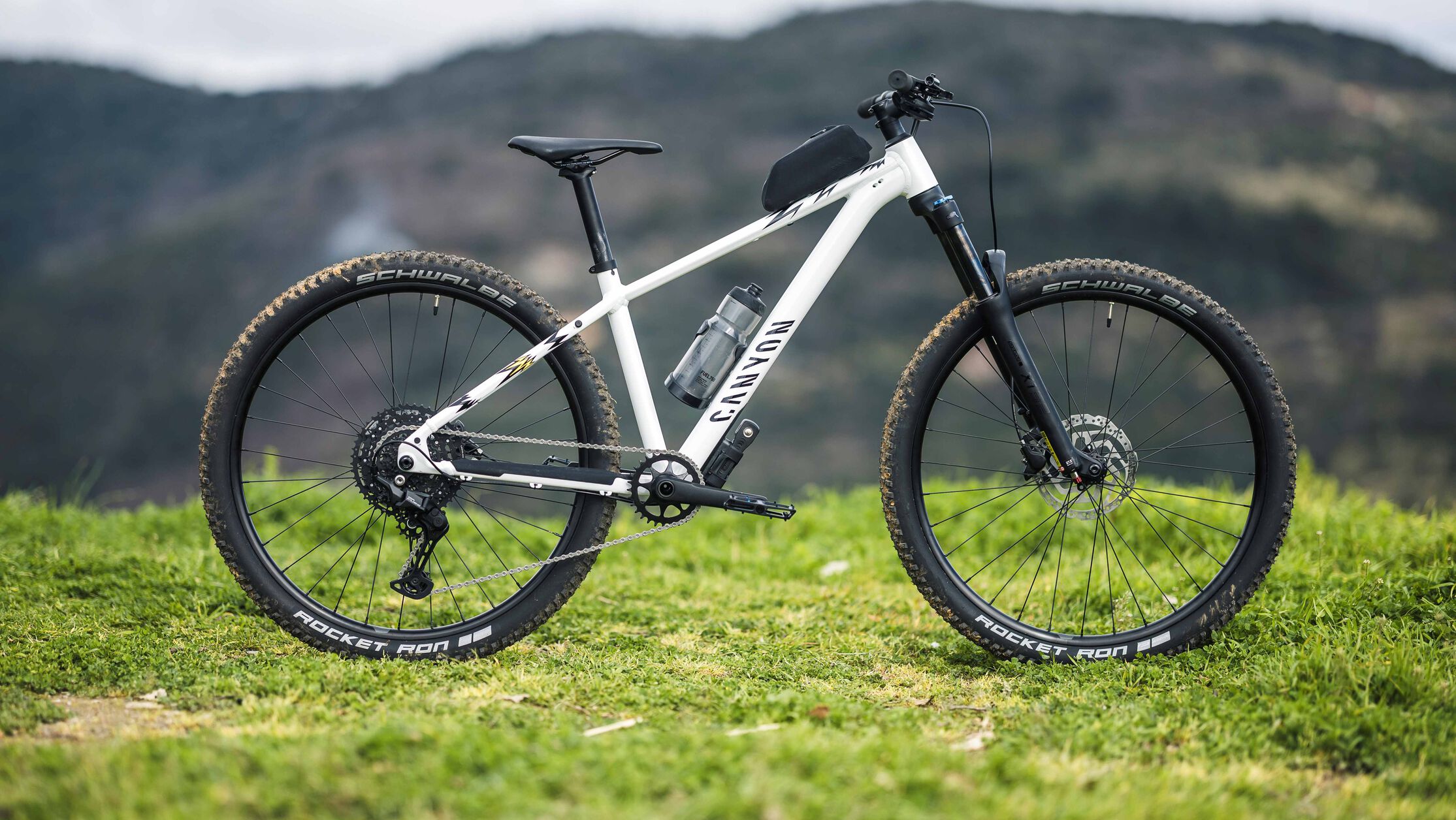 Side view of the Canyon Grand Canyon AL 8 hardtail mountain bike set on a grassy hill, featuring front suspension, Schwalbe Rocket Ron tires, and a lightweight aluminum frame.