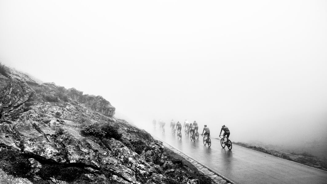 Cyclists riding through dense fog on a steep mountain road during La Vuelta a Espa&ntilde;a, surrounded by rocky terrain and low visibility.